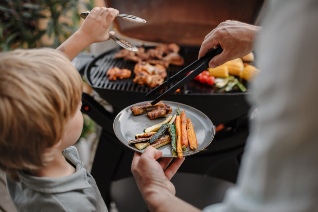 Gasgrill med mad på og lille dreng ved siden af.